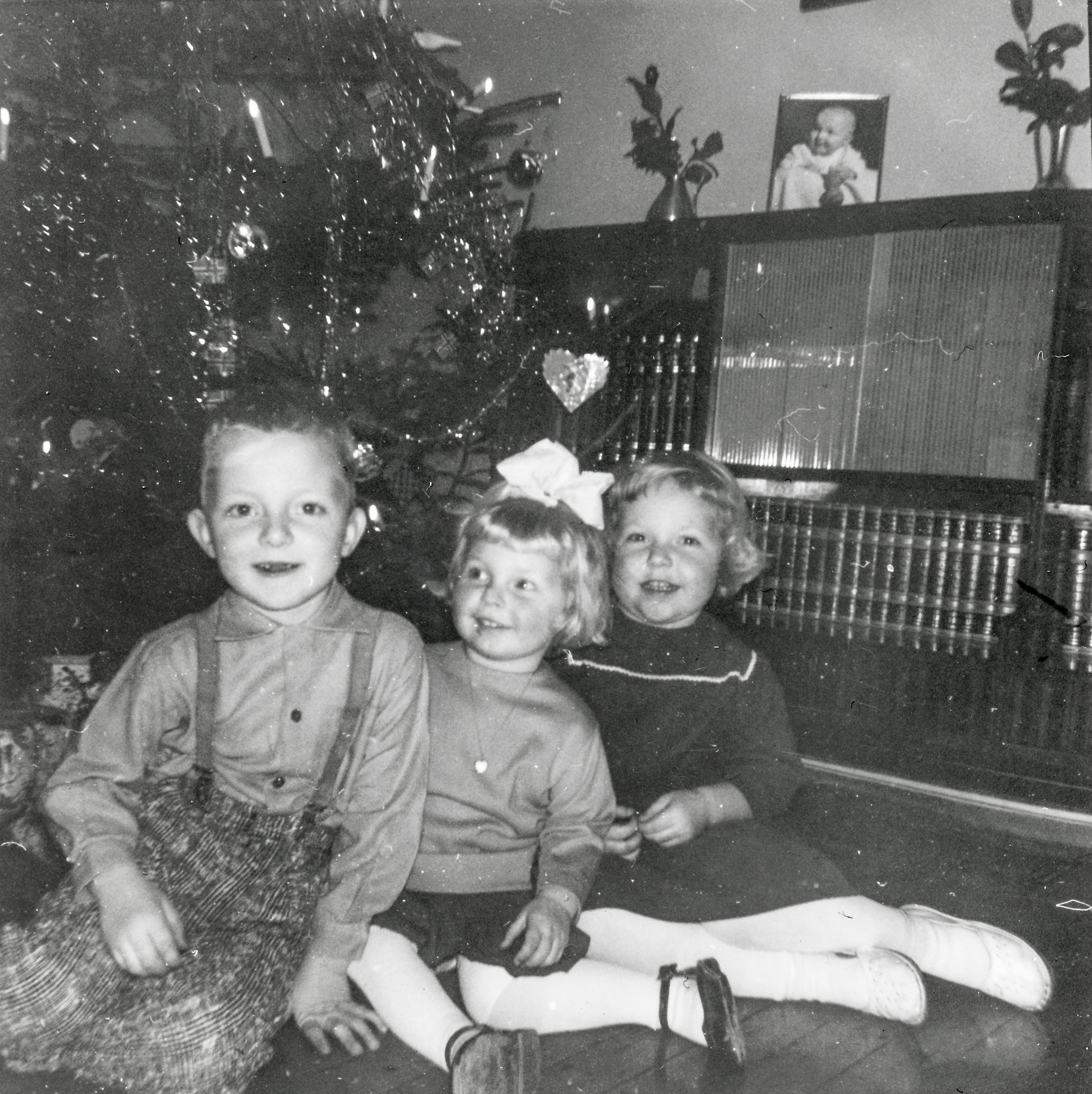 Tre barn sitter forventningsfulle foran et pyntet juletre med gaver under. Her har de elektriske juletrelysene gjort sitt inntog. Foto: Norsk Vasskraft- og Industristadmuseum