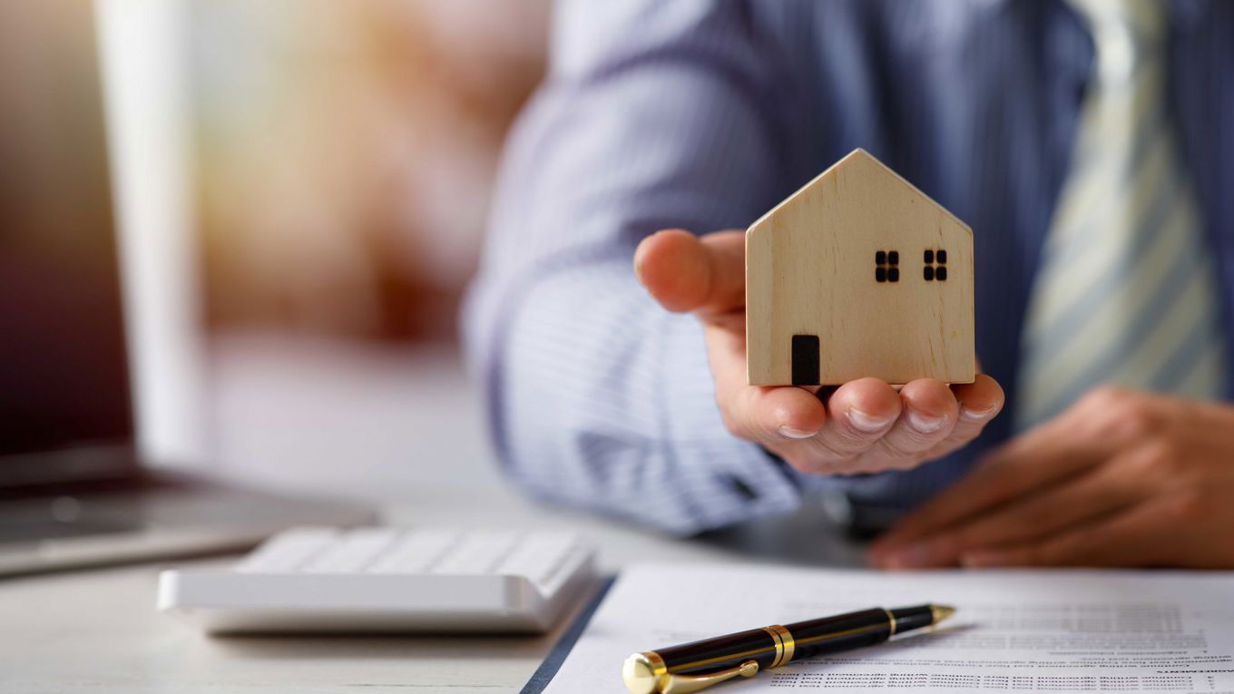 En mann holder et lite hus i hånden over et dokument og en penn