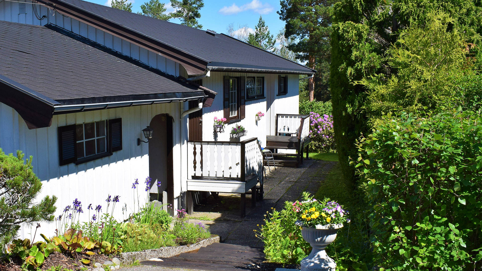 Vakkert hvitt bolighus med svarte takstein utvendig, front og inngang med grønn flott hage og blå himmel i sol- og sommersesongen i Norge