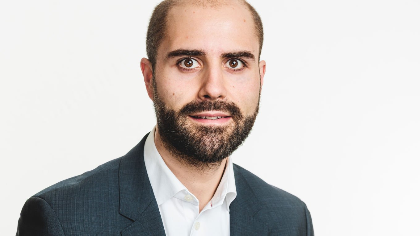 Mann med skjegg og kort hår i mørk dressjakke og hvit skjorte, ser mot kamera med nøytral bakgrunn.