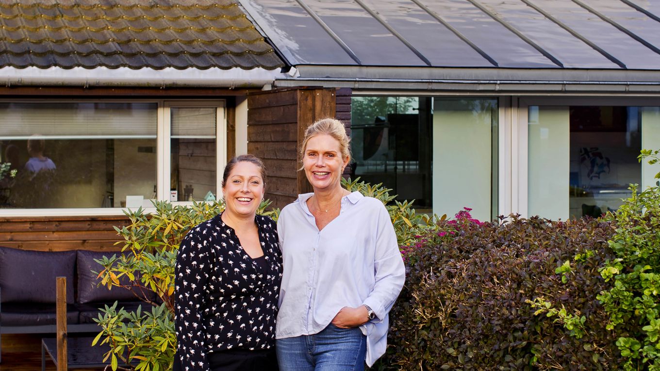 To kvinner står smilende foran en tomannsbolig med hage og terrasse.