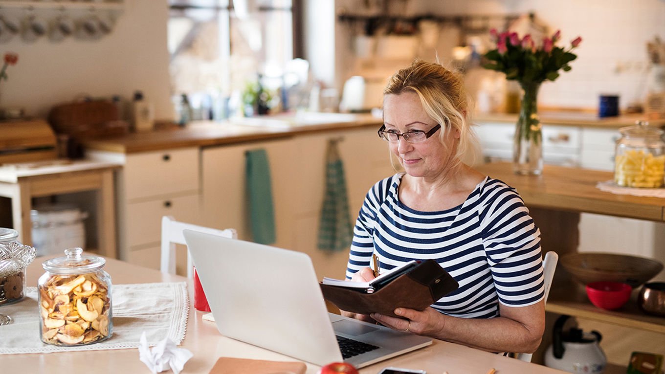 Dame sitter foran en bærbar PC på kjøkkenet og ser i almanakken sin