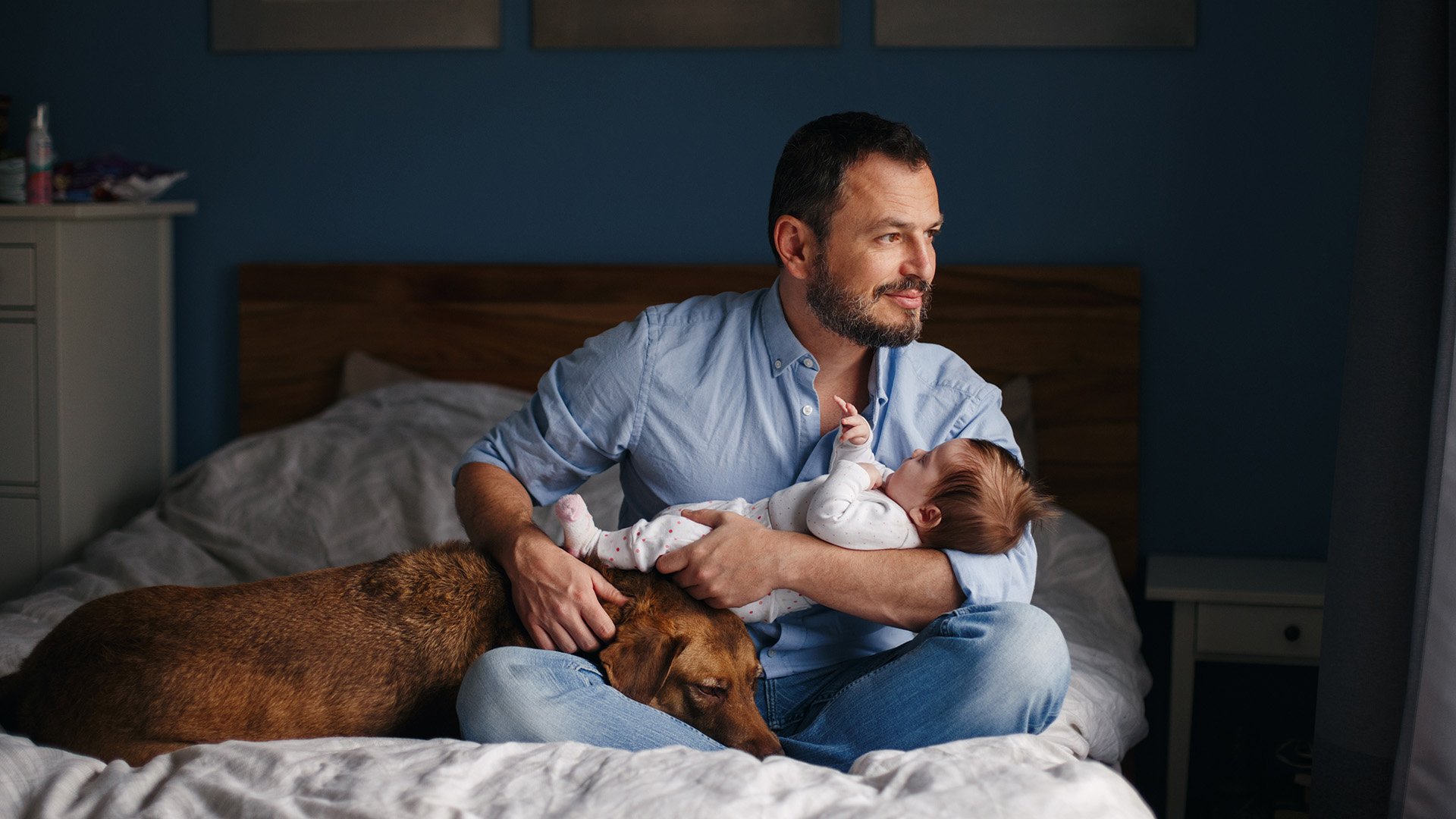 En mann i 30-årene sitter på en stor seng og har et spedbarn i armene. En hund ligger ved siden av ham på sengen og hviler hodet sitt i fanget til mannen. Mannen ser ut av vinduet.