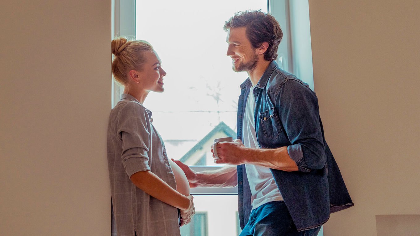 En mann og en høygravid kvinne i 30-årene står i en vindusåpning i en oppgang i et leilighetsbygg.