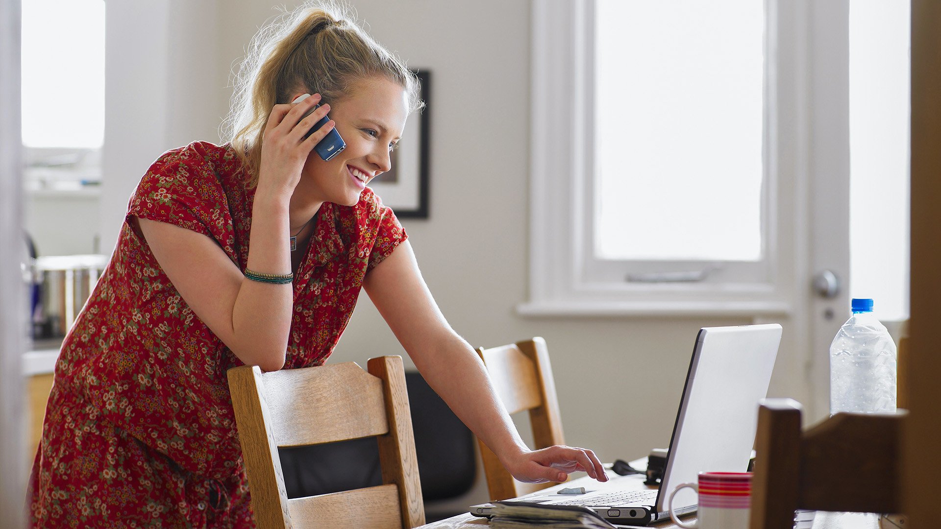 En ung kvinne står lent over en laptop som står på et kjøkkenbord mens hun snakker i telefonen.
