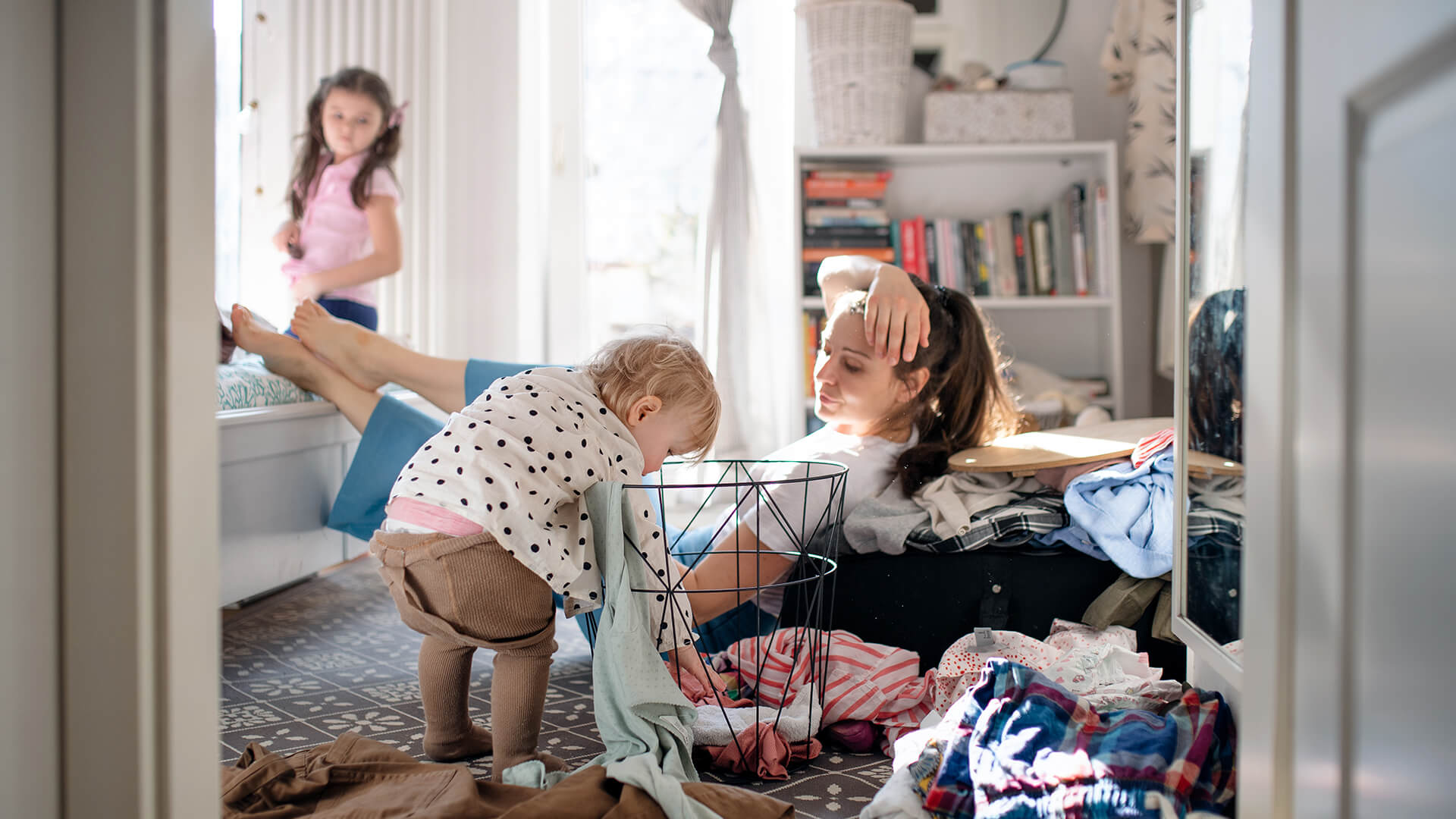 En kvinne i 30-årene sitter på soveromsgulvet og ser på mens et lite barn roter i en kleshaug. Et litt større barn står i bakgrunnen.