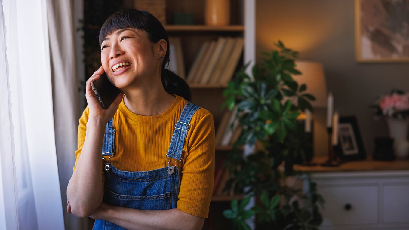 En smilende kvinne i 30-årene står ved et vindu i stuen sin og prater i telefonen.
