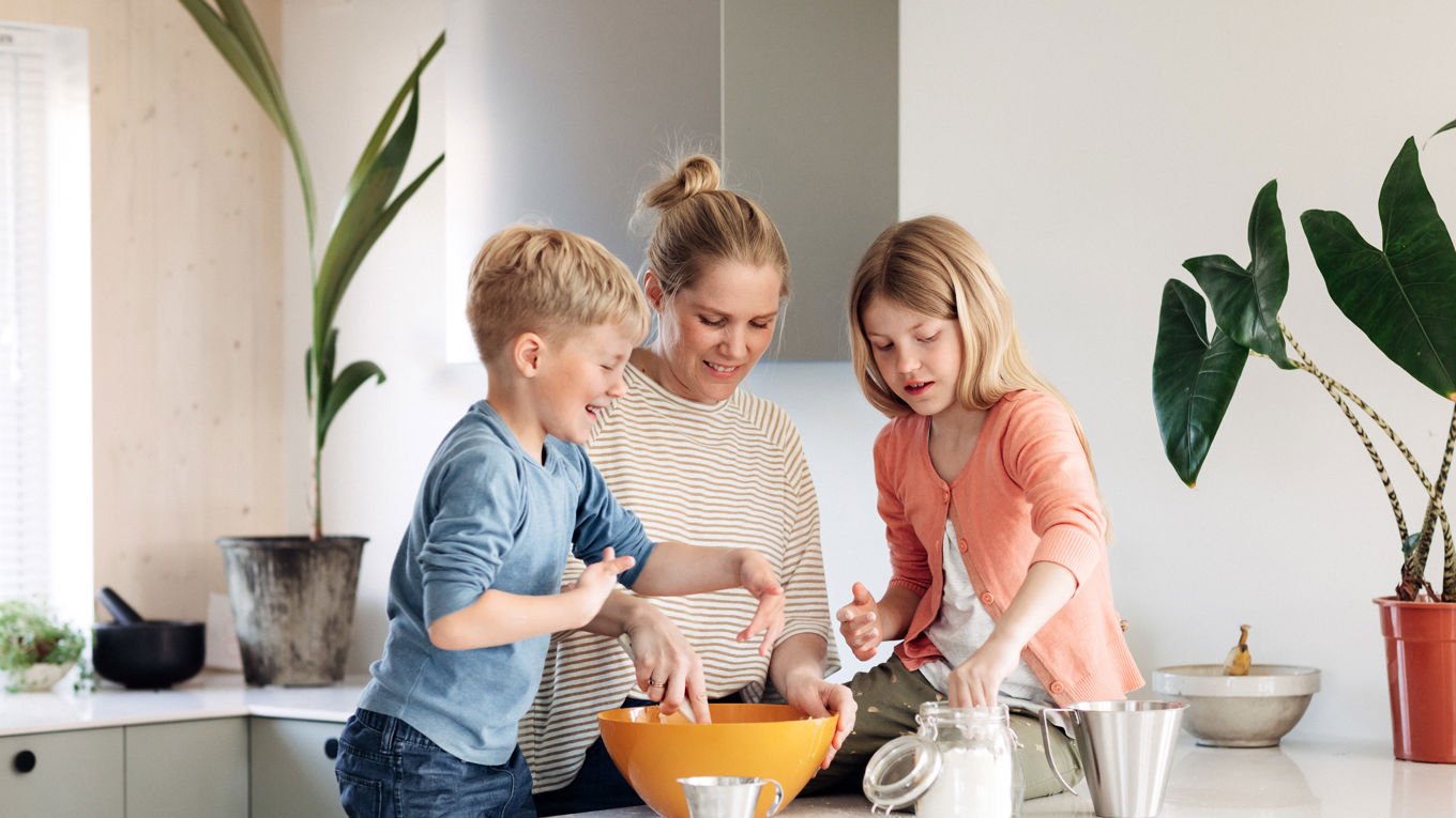 En dame i 40-årene og to barn står på kjøkkenet i et moderne hus. De samarbeider om å bake noe.