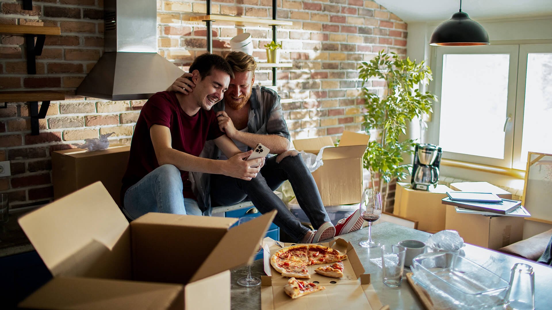 To unge menn sitter oppå kjøkkenbenken i en leilighet full av pappesker. De holder rundt hverandre og ser på mobilen. En take away-pizza og vinglass står på bordet.
