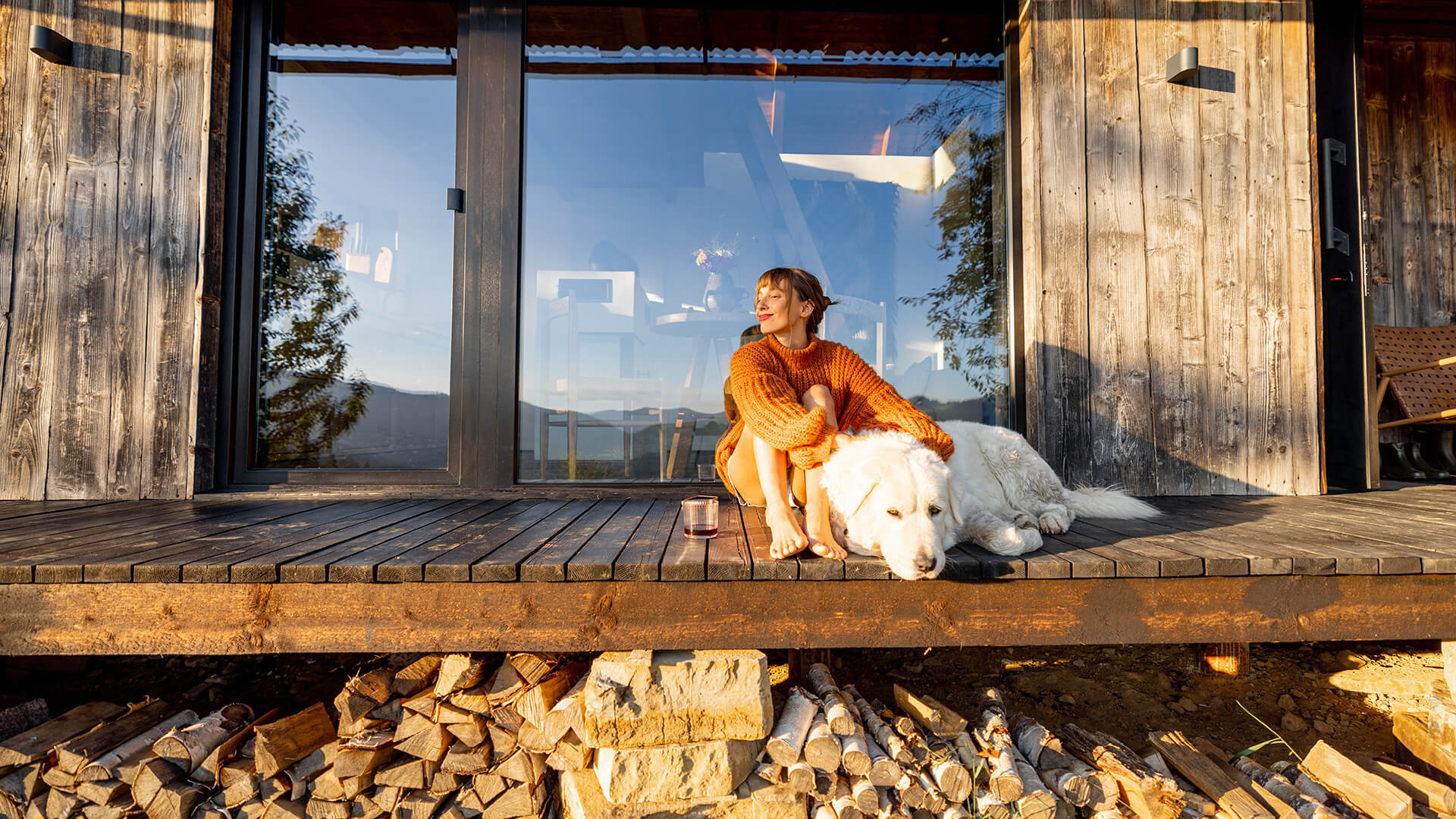 En ung kvinne sitter på en terrasse av tre sammen med en stor hund. Bak henne er en skyvedør av glass og værbitt trepanel. Under terrassen er det stablet masse vedkubber.