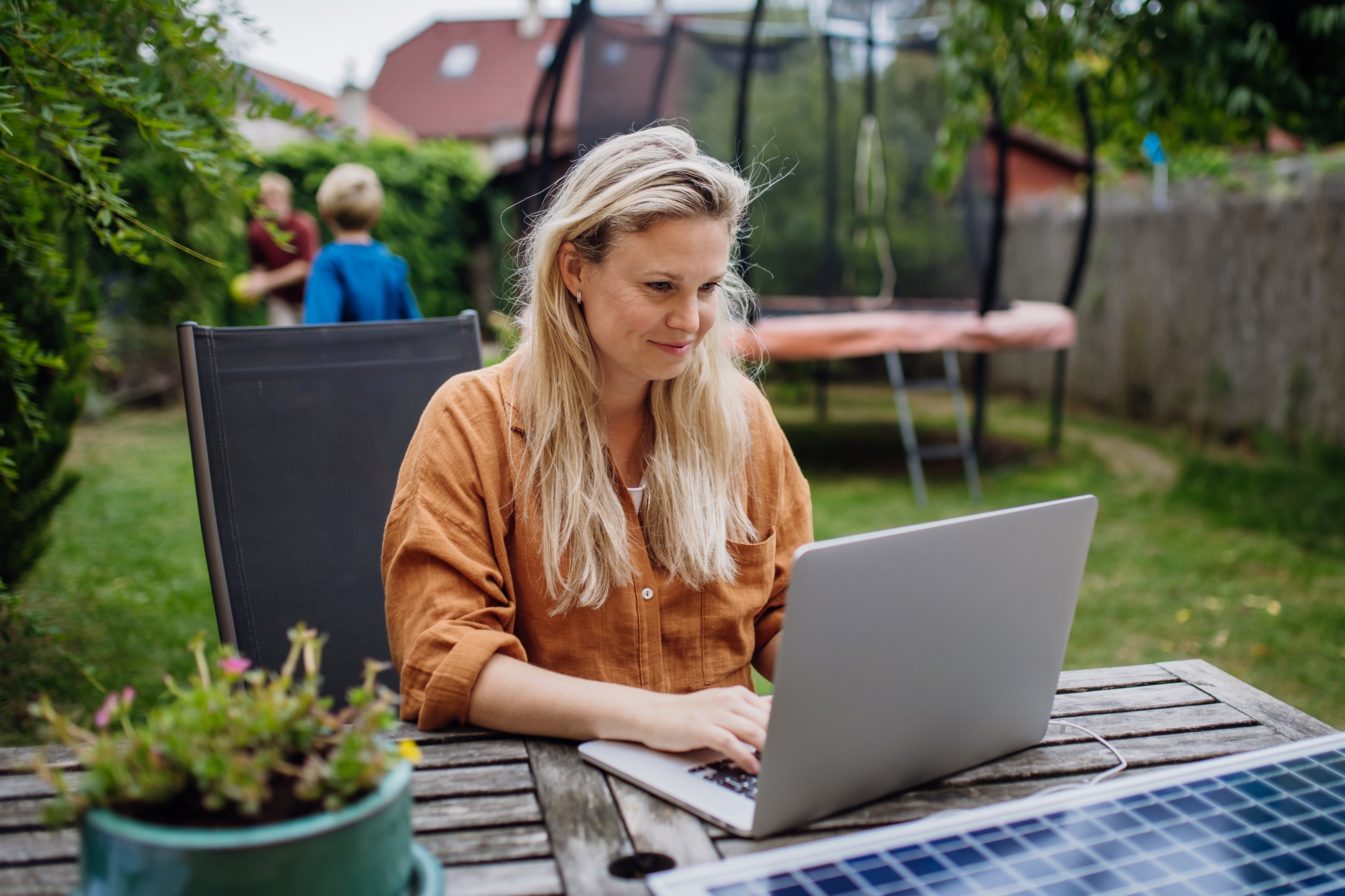 En kvinne i 40-årene sitter ved et bord ute i hagen og tar et digitalt kurs mens to barn leker. En trampoline og flere hus synes i bakgrunnen.