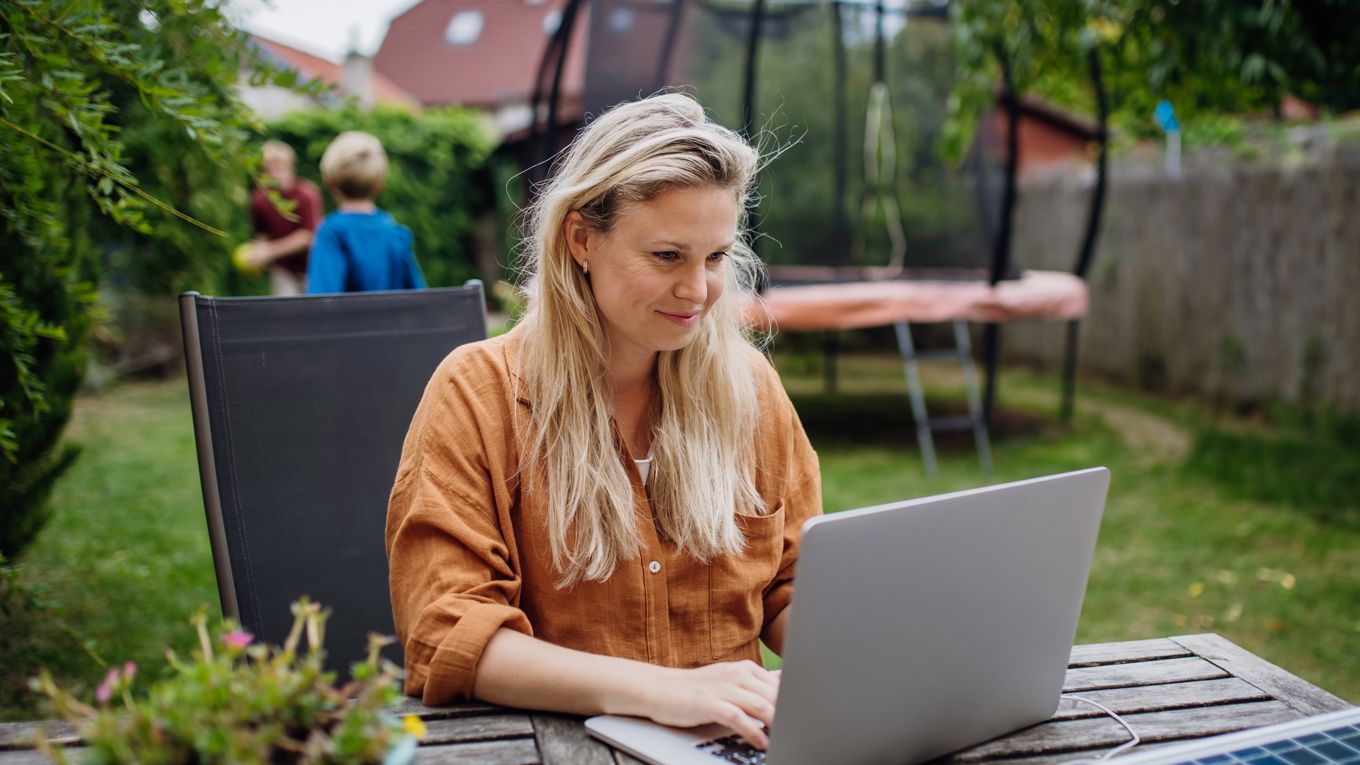 En kvinne i 40-årene sitter ved et bord ute i hagen og tar et digitalt kurs mens to barn leker. En trampoline og flere hus synes i bakgrunnen.