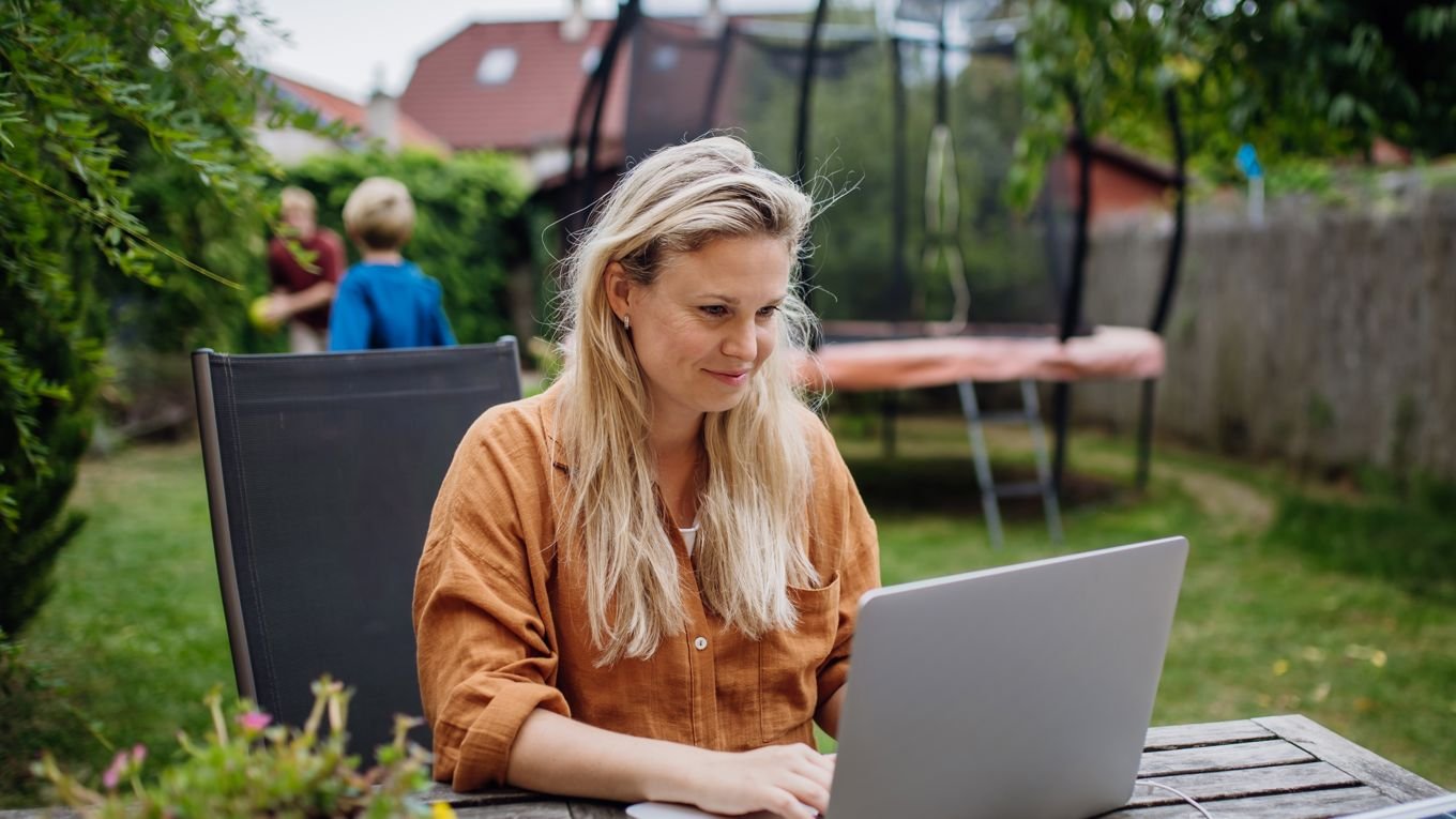 En kvinne i 40-årene sitter ved et bord ute i hagen og tar et digitalt kurs mens to barn leker. En trampoline og flere hus synes i bakgrunnen.