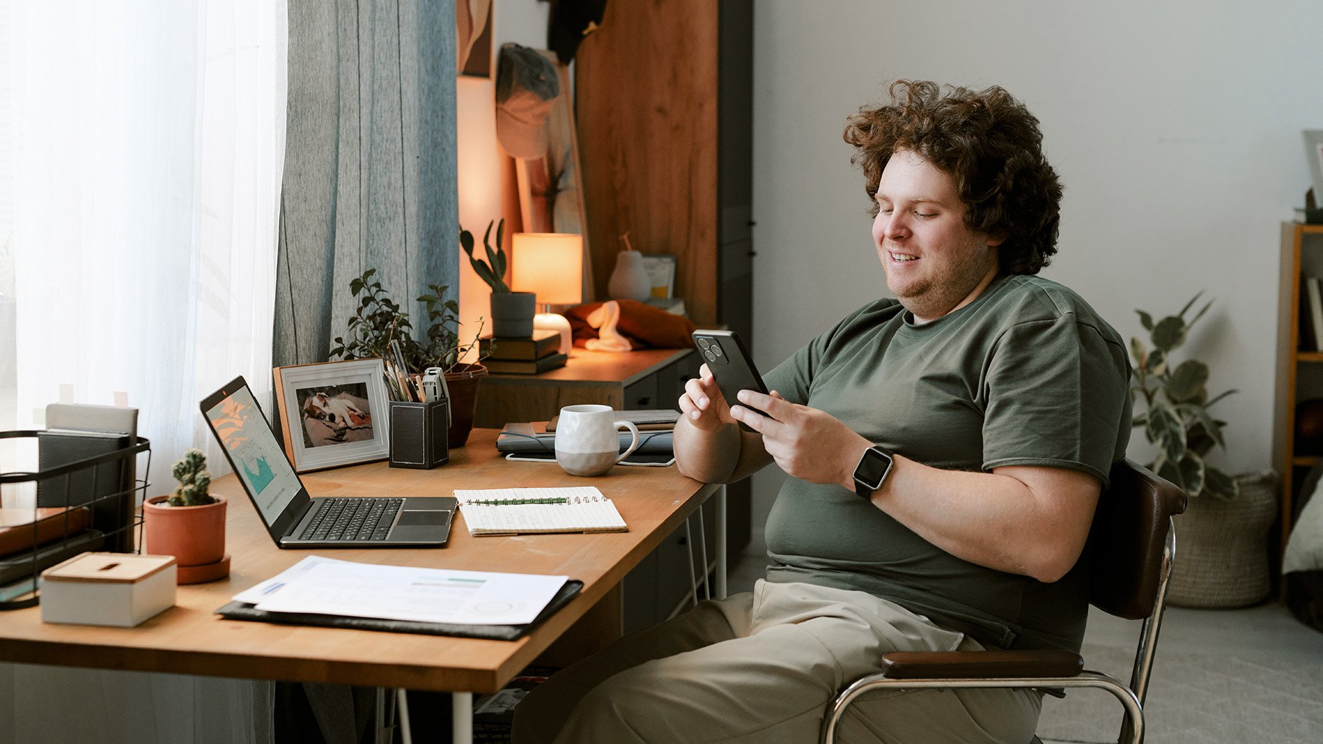 En mann i 30-årene sitter ved en pult på hjemmekontor og ser på mobilen mens han smiler. Foran ham på pulten står en laptop og en rekke bøker og papirer.