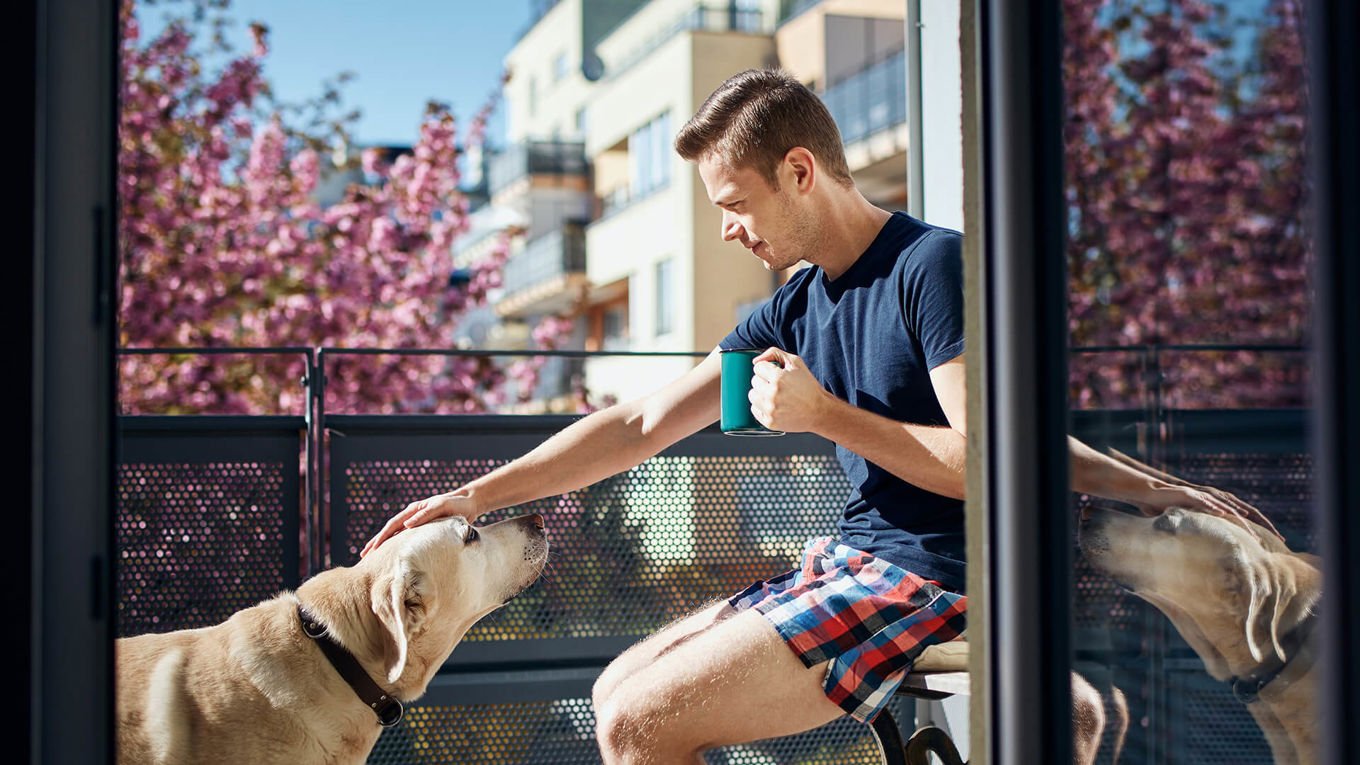 En ung mann sitter på balkongen i leiligheten sin og klapper en hund på hodet. Han har sommerklær på og kaffekopp i hånden. I bakgrunnen ses et tre med rosa blomster og flere leilighetsbygg.