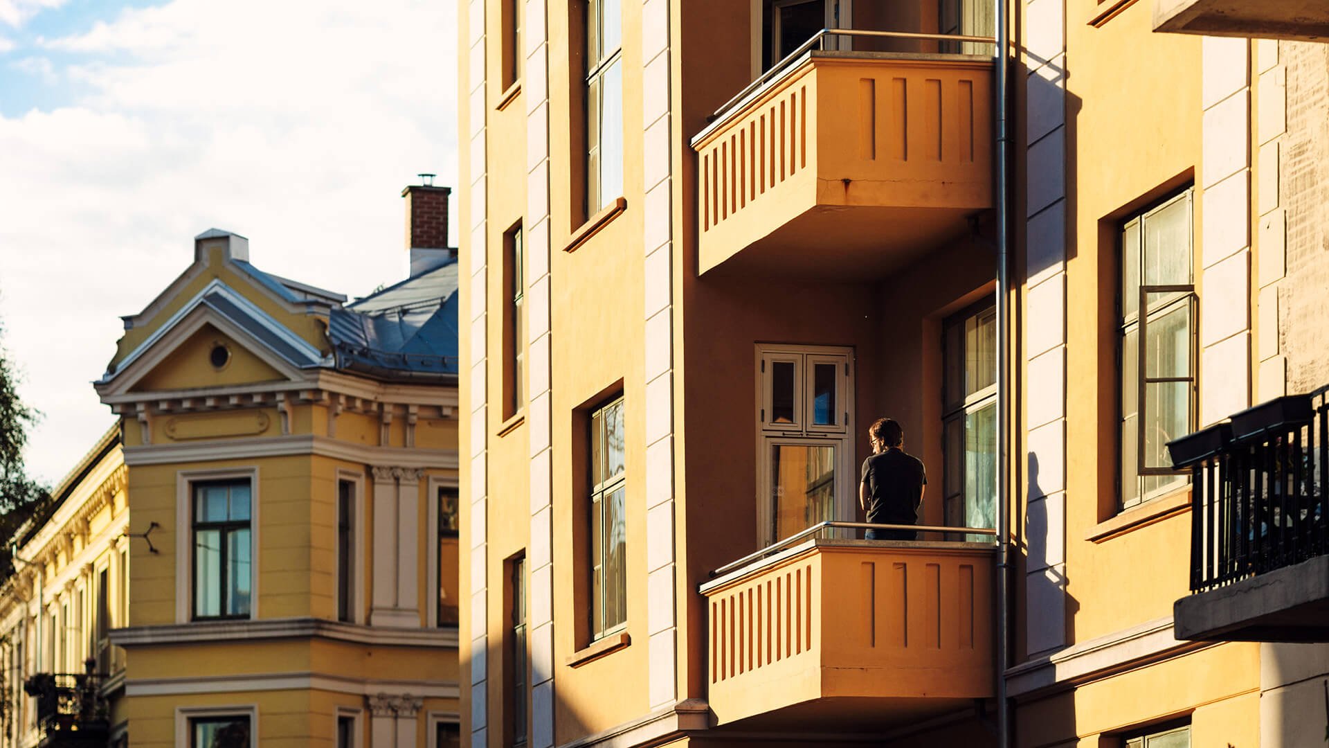 Fasaden til en klassisk gul bygård med balkonger. På den ene balkongen står en mann med ryggen mot kamera.