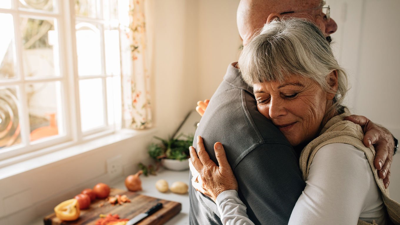 En kvinne i 60-årene omfavner en jevnaldrende mann. I bakgrunnen syns en kjøkkenbenk og litt oppkuttede grønnsaker.