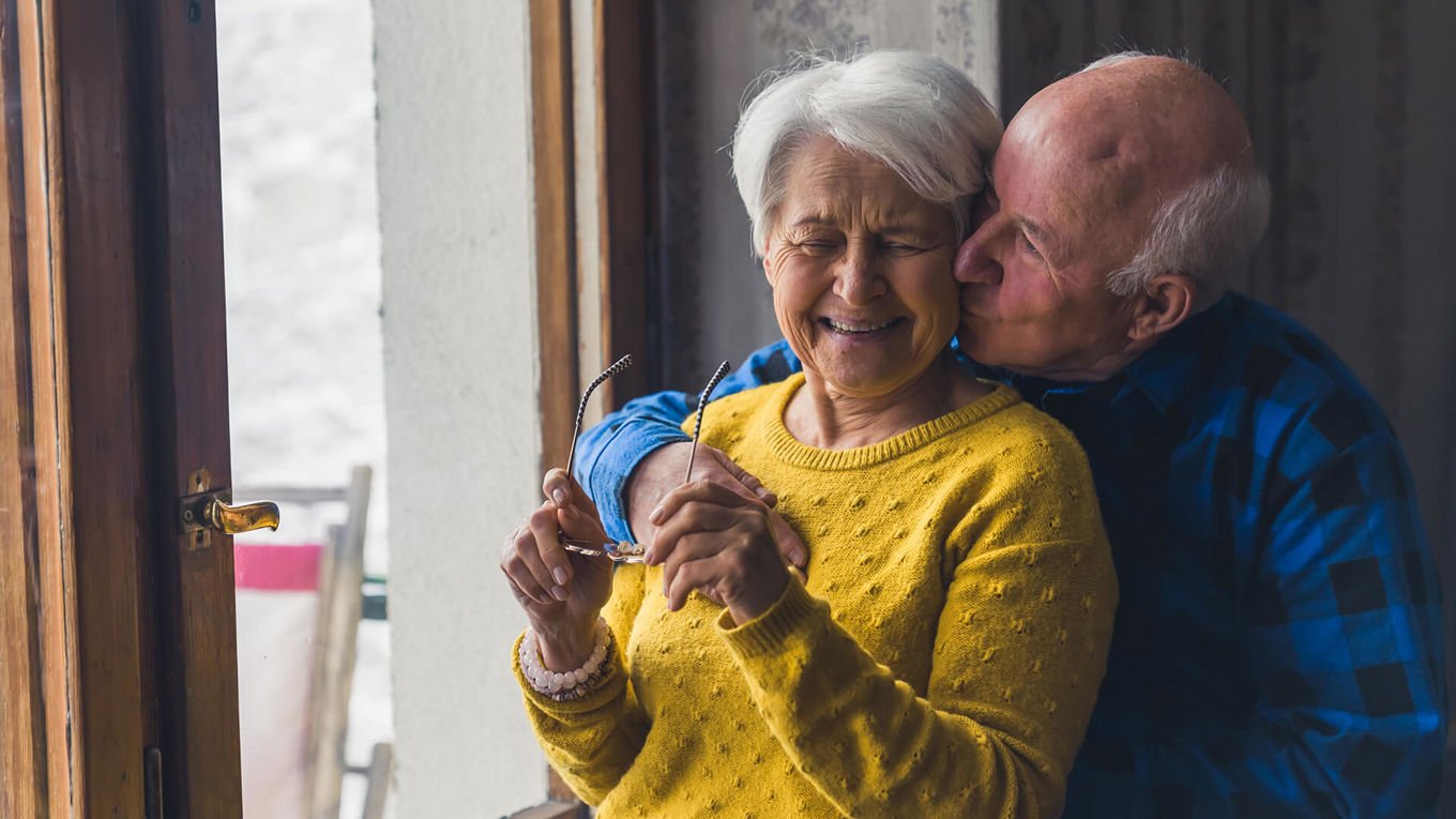 Et ektepar i 70-årene står ved et vindu i et hus og klemmer hverandre. Mannen gir kvinnen et kyss på kinnet, og kvinner ler.