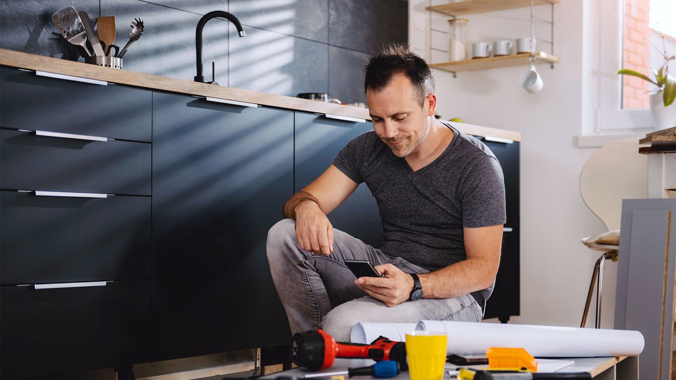 En mann sitter på gulvet i et moderne kjøkken og ser på mobilen sin. Foran ham ligger verktøy, en gul kopp og tegninger, som tyder på at han holder på med et oppussings- eller monteringsprosjekt. Lyset strømmer inn gjennom et vindu og kaster skygger på veggen.