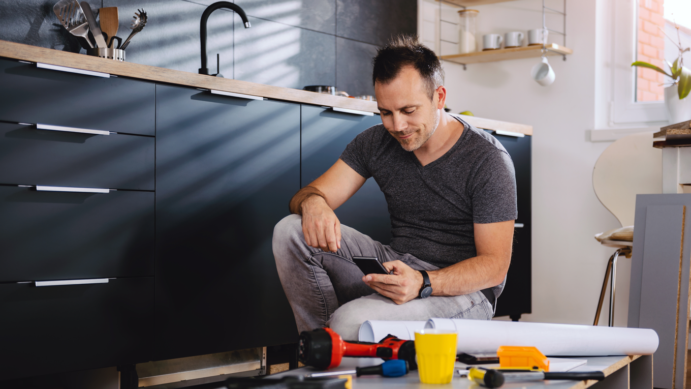 En mann sitter på gulvet i et moderne kjøkken og ser på mobilen sin. Foran ham ligger verktøy, en gul kopp og tegninger, som tyder på at han holder på med et oppussings- eller monteringsprosjekt. Lyset strømmer inn gjennom et vindu og kaster skygger på veggen.