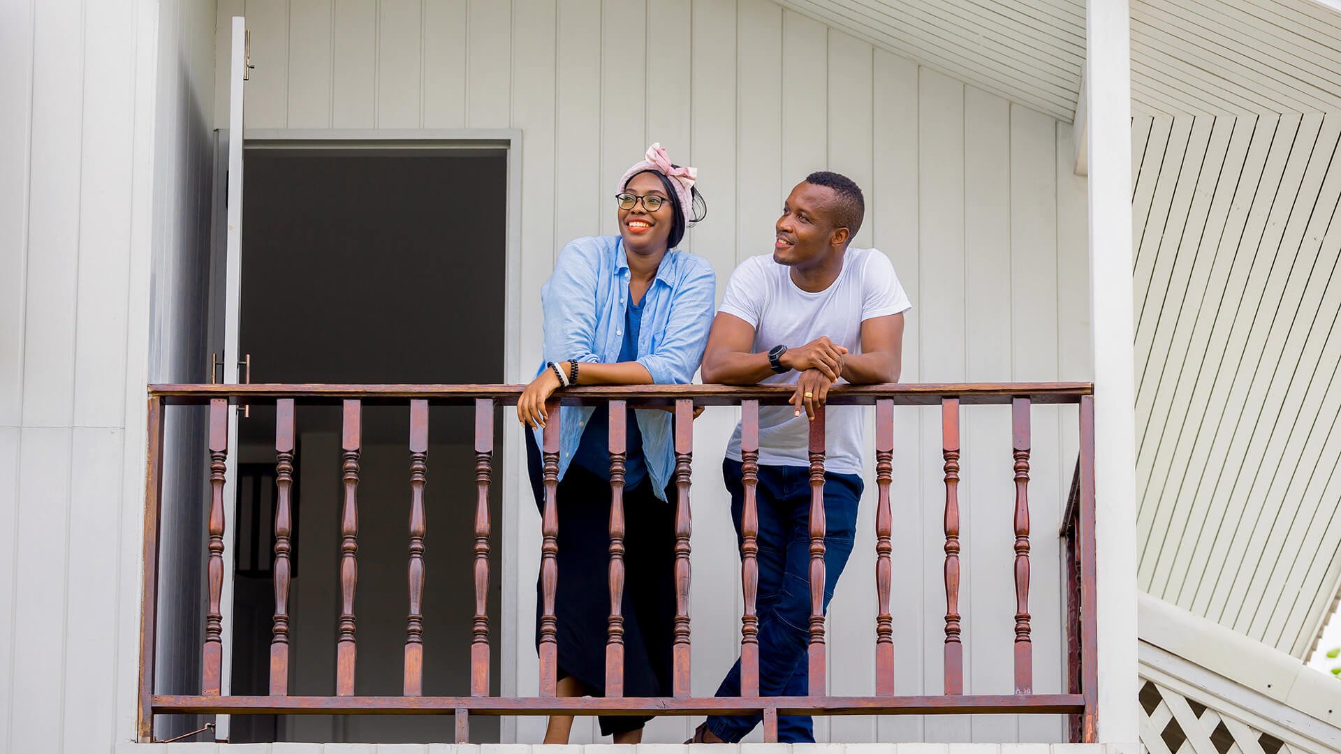 Et ungt par står på en balkong med trerekkverk utenfor en hvitt trehus.