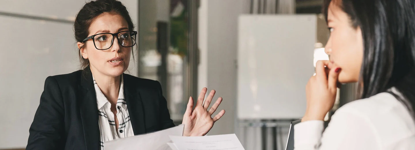 To personer sitter ved et bord og snakker sammen, begge holder papirer i hånden