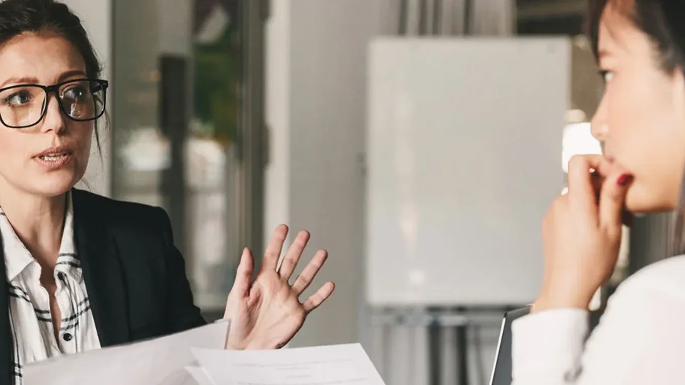 To personer sitter ved et bord og snakker sammen, begge holder papirer i hånden