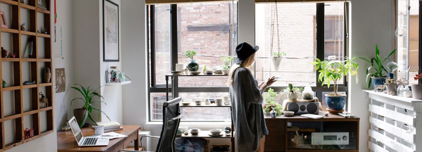 En person med hatt står i en lys leilighet med mange planter og bruker mobilen ved vinduet.