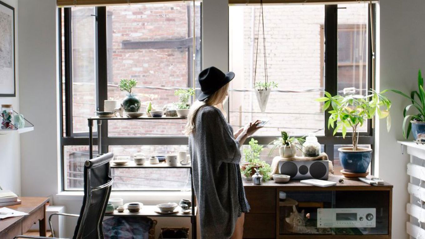 En person med hatt står i en lys leilighet med mange planter og bruker mobilen ved vinduet.